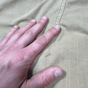May include: A tan-colored fabric garment featuring a subtle grid-like texture and a visible vertical seam. The material displays a light, neutral tone with a small, faint circular discoloration mark located near the center of the visible surface area.