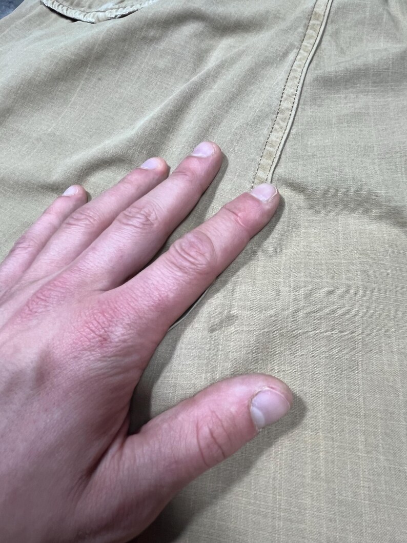 May include: A tan-colored fabric garment featuring a subtle grid-like texture and a visible vertical seam. The material displays a light, neutral tone with a small, faint circular discoloration mark located near the center of the visible surface area.