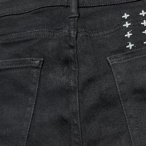 Op de afbeelding: Close-up van de achterkant van een zwarte spijkerbroek. Witte kruisborduurwerk is zichtbaar op de rechter achterzak. De jeans heeft een klassiek ontwerp met vijf zakken met een knoop- en ritssluiting.