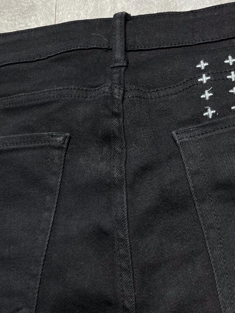 Op de afbeelding: Close-up van de achterkant van een zwarte spijkerbroek. Witte kruisborduurwerk is zichtbaar op de rechter achterzak. De jeans heeft een klassiek ontwerp met vijf zakken met een knoop- en ritssluiting.