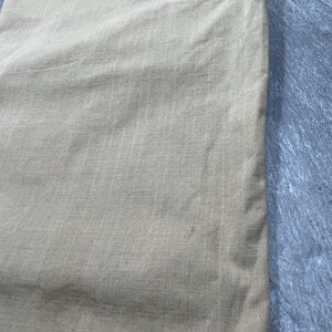May include: A pair of light tan trousers featuring a subtle grid-like ripstop texture. The fabric has a visible weave pattern with a small, faint discoloration mark on the lower section. The garment includes a stitched seam detail at the top.
