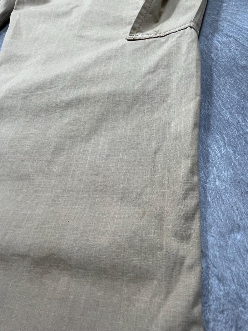 May include: A pair of light tan trousers featuring a subtle grid-like ripstop texture. The fabric has a visible weave pattern with a small, faint discoloration mark on the lower section. The garment includes a stitched seam detail at the top.