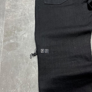Op de afbeelding: Close-up van een zwarte denim jeans. De jeans heeft een klein, zilverkleurig logo bij de zoom en een gerafeld detail. De stof is donker gewassen, met een klassiek ontwerp met vijf zakken.