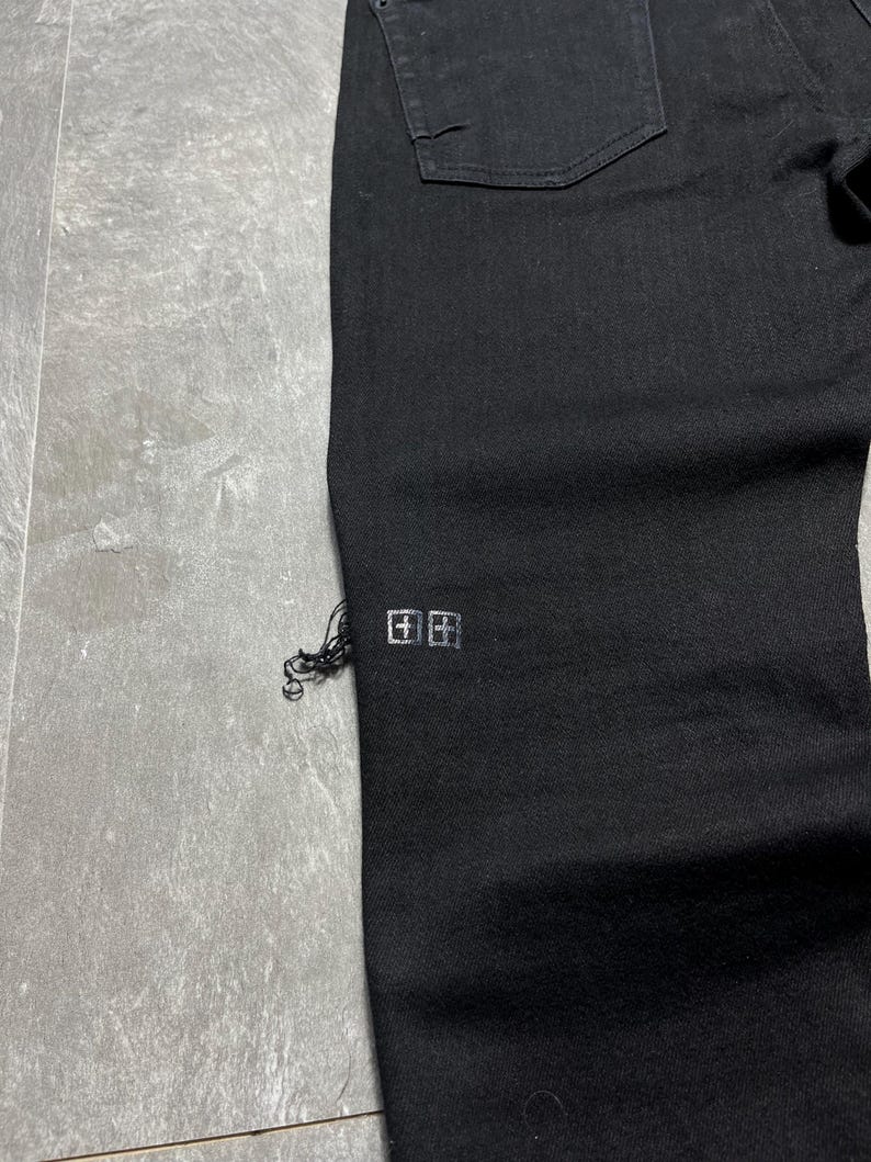 Op de afbeelding: Close-up van een zwarte denim jeans. De jeans heeft een klein, zilverkleurig logo bij de zoom en een gerafeld detail. De stof is donker gewassen, met een klassiek ontwerp met vijf zakken.