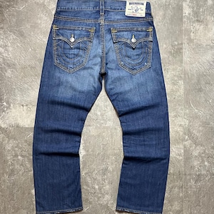 May include: Blue denim jeans with a faded wash. The back pockets have a decorative stitching design. The jeans have a True Religion label on the back. The stitching is yellow.