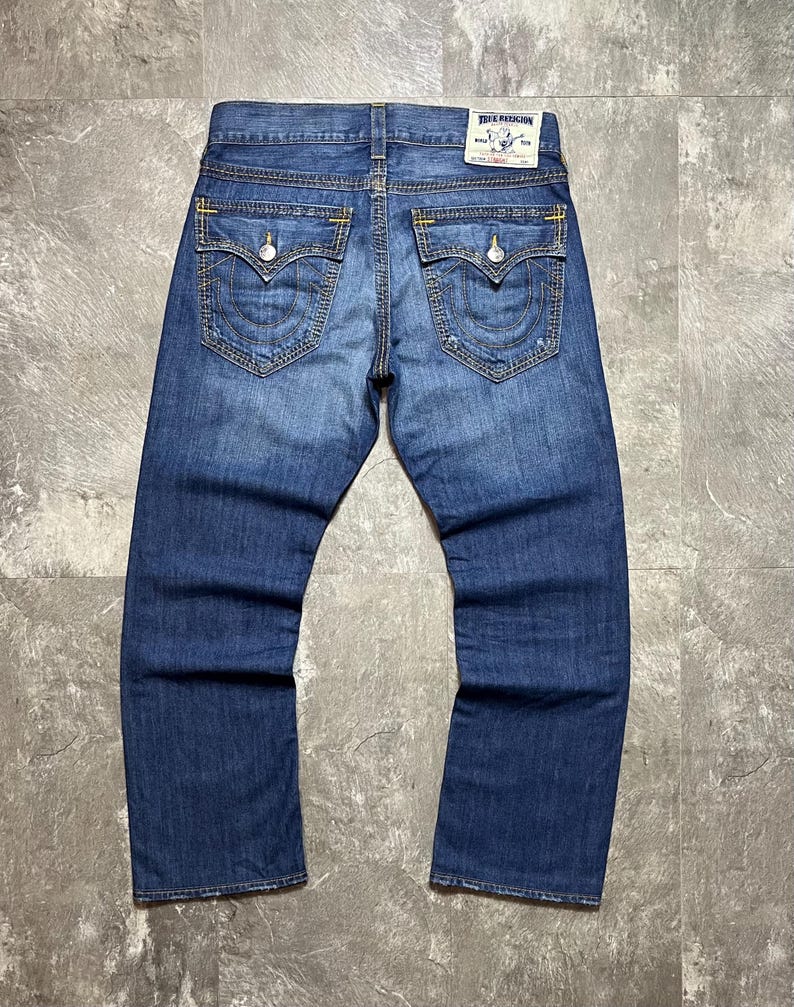 May include: Blue denim jeans with a faded wash. The back pockets have a decorative stitching design. The jeans have a True Religion label on the back. The stitching is yellow.