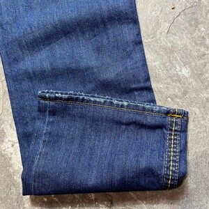 May include: Close-up of blue denim jeans. The jeans are folded at the hem, revealing yellow stitching. The fabric has a textured appearance, and the overall color is a deep indigo.