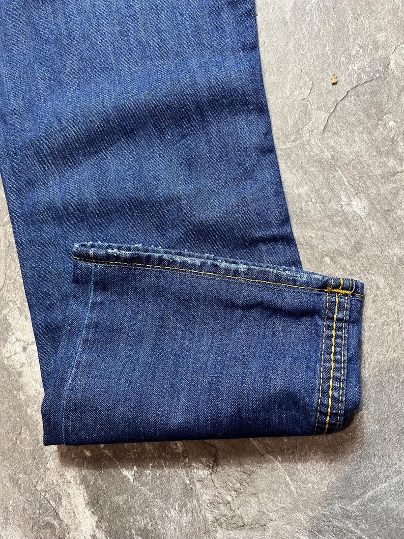 May include: Close-up of blue denim jeans. The jeans are folded at the hem, revealing yellow stitching. The fabric has a textured appearance, and the overall color is a deep indigo.