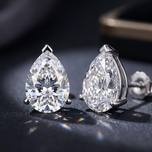 Può includere: Un paio di orecchini a lobo con pietre trasparenti sfaccettate a forma di pera, incastonate in metallo color argento lucido. Ogni orecchino è fissato con un perno e una chiusura abbinata, mostrando una superficie brillante e riflettente.