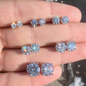 May include: Three pairs of round, clear, faceted stud earrings arranged in a vertical row. Each earring features a single brilliant-cut stone set in a metallic prong setting, reflecting light with a sparkling, multi-colored prismatic effect.