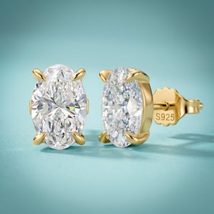 May include: A pair of gold-colored stud earrings with oval-shaped, clear gemstones. The earrings have a post and backing. The metal is stamped with "S925". The background is a solid light blue.