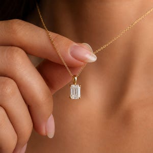 May include: A gold chain necklace featuring a single emerald-cut clear gemstone pendant held in a four-prong setting. The pendant hangs from a delicate gold link chain, showcasing a rectangular faceted stone that reflects light with clarity.