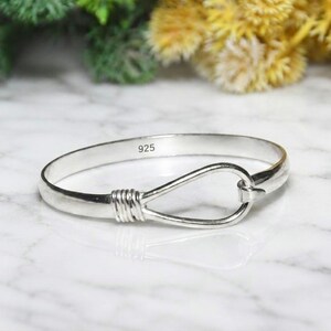 May include: A polished silver bangle bracelet featuring a hook-and-eye style closure with a wrapped wire detail. The interior of the band is clearly stamped with the hallmark 925, indicating sterling silver composition. The metal has a smooth finish.