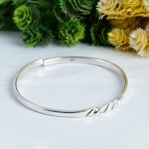 May include: A polished silver bangle bracelet. The bracelet has a smooth finish and a decorative wave pattern on one side. The number "925" is stamped inside, indicating the silver content. The bangle is a classic piece of jewellery.