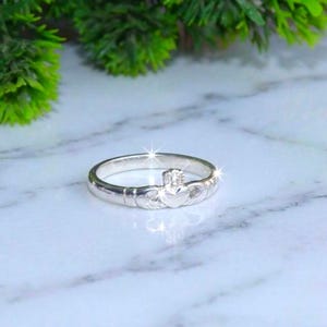 May include: A silver Claddagh ring featuring a central heart topped with a crown and flanked by two hands. The band is polished and reflective, showcasing the traditional design elements of the heart, crown, and hands in a metallic silver finish.