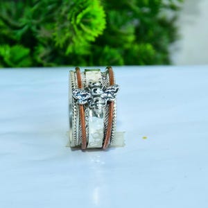 May include: A silver ring with a bee design. The ring features a central band with a textured pattern, flanked by copper-colored bands. The bee is silver with detailed wings. The ring is set against a light background.