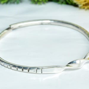 May include: A silver-toned bangle bracelet with a twisted design. One section of the bracelet features a series of rectangular indentations. The bracelet is displayed on a white surface, with greenery in the background.