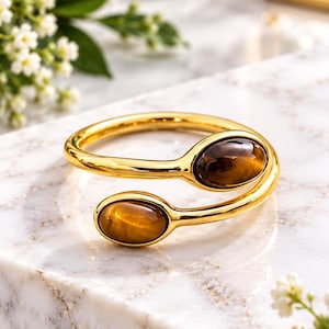 Puede incluir: Un brazalete abierto de tono dorado con dos piedras preciosas de ojo de tigre de forma ovalada fijadas en cada extremo. La banda de metal pulido se curva para crear un diseño flexible y abierto, resaltando la piedra marrón y dorada.