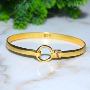 May include: A gold-toned bangle bracelet featuring a circular clasp mechanism and a ribbed detail on one side. The interior of the band is clearly stamped with the text 925, indicating the metal composition of the jewelry piece.