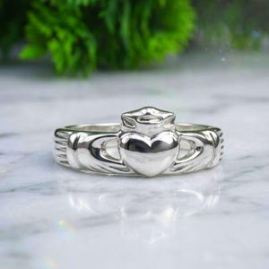 May include: A polished silver Claddagh ring featuring a central heart topped with a crown, held by two hands on either side of the band. The metal surface is smooth and reflective, showcasing the traditional design elements of the ring clearly.