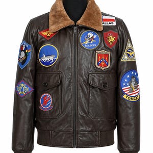 May include: Brown leather bomber jacket with various embroidered patches. It has a brown faux fur collar, two front pockets, and ribbed hem and cuffs. Patches include text such as "TOP TEN" and "TOMCAT".