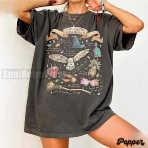 May include: A charcoal gray short-sleeve t-shirt featuring a graphic print titled Wizard Essentials. The design includes a wizard hat, an owl carrying a letter, a potion bottle, a mandrake, a broomstick, and glasses, surrounded by small stars.