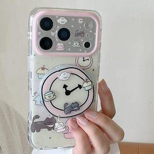 May include: Clear phone case with a pink border and a pop-up grip featuring a clock design. The case has a whimsical pattern of cats, cupcakes, and coffee cups. The phone case is held by a person.