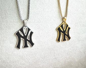 Könnte beinhalten: Zwei Halsketten mit dem New York Yankees Logo als Anhänger. Eine Kette hat eine silberfarbene Kette und einen Anhänger, die andere eine goldfarbene Kette und einen Anhänger. Beide Anhänger sind mit einer schwarzen Emaille-Einlage versehen.