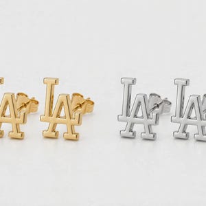 May include: Two pairs of stud earrings featuring the letters LA in a stylised font. One pair is finished in a polished gold tone, while the other pair is finished in a polished silver tone. Each earring includes a standard post and butterfly back.