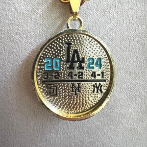 以下が含まれることがあります： 金色のペンダントネックレス。円形のチャームには、黒色の「LA」の文字と、青色の数字「20」と「24」が表示されています。その下に野球の試合のスコアがあります。ネックレスは金色のチェーンです。