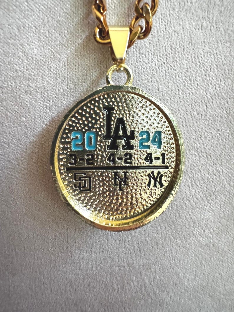 以下が含まれることがあります： 金色のペンダントネックレス。円形のチャームには、黒色の「LA」の文字と、青色の数字「20」と「24」が表示されています。その下に野球の試合のスコアがあります。ネックレスは金色のチェーンです。