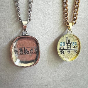 以下が含まれることがあります： 2つのペンダントネックレス。1つはシルバーチェーンと「LA」の文字と数字が入った銅色のペンダント。もう1つはゴールドチェーンと「LA」の文字と数字が入ったゴールドのペンダント。どちらのペンダントもスポーツをテーマにしています。
