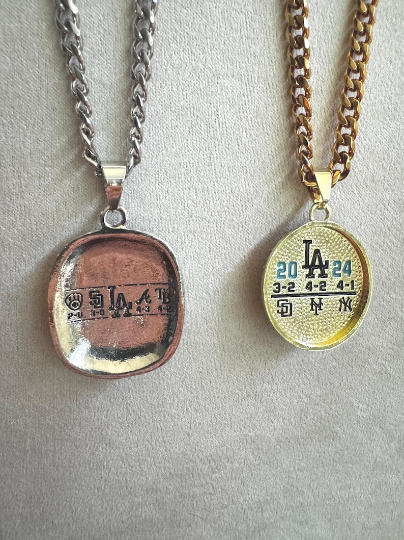 以下が含まれることがあります： 2つのペンダントネックレス。1つはシルバーチェーンと「LA」の文字と数字が入った銅色のペンダント。もう1つはゴールドチェーンと「LA」の文字と数字が入ったゴールドのペンダント。どちらのペンダントもスポーツをテーマにしています。