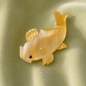 May include: A translucent golden-yellow fish-shaped brooch with a smooth, polished finish and a single small black circular eye. The accessory features detailed etched lines along the fins and tail, creating a textured appearance on the surface.