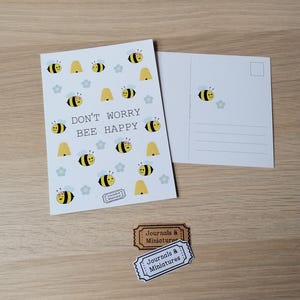 Könnte beinhalten: Zwei weiße Postkarten mit Bienen-Illustrationen und dem Text "DON'T WORRY BEE HAPPY". Eine Karte zeigt die Vorderseite mit Bienen, Blumen und einem Bienenstock. Die andere zeigt die Rückseite mit einer einzelnen Biene und einem Stempelfeld. Zwei kleine Etiketten sind ebenfalls vorhanden.
