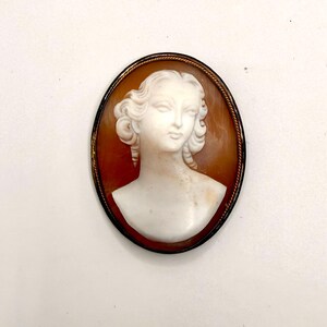 Op de afbeelding: Een ovale cameebroche met een wit uitgesneden profiel van een persoon met golvend haar tegen een doorschijnende amberkleurige achtergrond. Het sieraad is gevat in een dunne, donkere metalen rand met een gedraaide touwstructuur langs de rand.