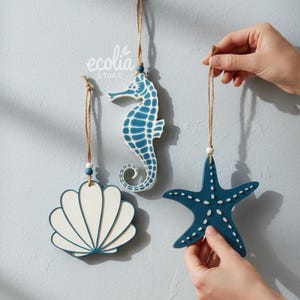 Peut inclure: Trois ornements côtiers suspendus : un coquillage blanc et bleu, un hippocampe bleu et une étoile de mer bleue. Chaque ornement est suspendu par une ficelle naturelle. L'hippocampe et l'étoile de mer ont des détails de points blancs.