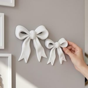 以下が含まれることがあります： 長いリボンが付いた2つの白い壁掛け用リボン装飾。リボンは滑らかでマットな質感で、中央に結び目があるクラシックなデザインです。これらの装飾要素は、ニュートラルな色の壁面に配置されています。