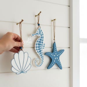 Peut inclure: Trois ornements suspendus sur le thème côtier : un coquillage, un hippocampe et une étoile de mer. Les ornements sont peints dans des tons de bleu et de blanc, avec une corde pour les suspendre. Un main tient le coquillage.