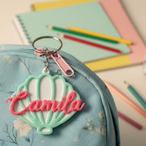 Puede incluir: Un llavero personalizado con el nombre "Camila" en escritura rosa, sujeto a una mochila azul claro. El llavero tiene un diseño festoneado verde y blanco. Lápices y cuadernos están en el fondo.