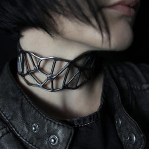 May include: A dark metallic choker necklace featuring an openwork geometric lattice design. The structure consists of interconnected angular segments forming a web-like pattern that encircles the neck, finished with a matte, slightly weathered texture.