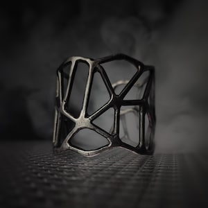 Può includere: Un ampio bracciale a fascia in metallo grigio scuro caratterizzato da un design a reticolo geometrico traforato. La struttura è composta da forme poligonali irregolari interconnesse con una finitura opaca e testurizzata, simile a una gabbia.