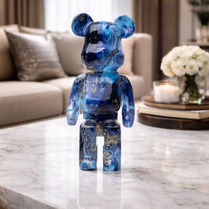 May include: A blue and gold decorative bear figurine. The figurine has a marbled appearance with shades of blue and gold accents. It is standing on a white marble table with a gold frame. The bear has a glossy finish and is a modern art piece.