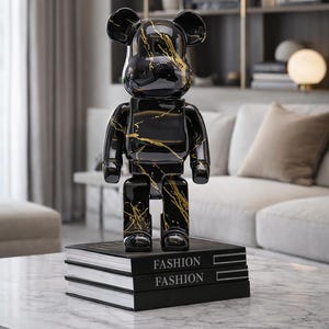 May include: A black and gold decorative bear figurine with a glossy finish, standing on a stack of black books labeled "FASHION". The bear has gold veining throughout its body. The figurine is on a marble coffee table.