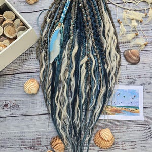 Op de afbeelding: Handgemaakte dreadlocks in tinten blauw, grijs en crème. De dreads zijn versierd met kralen en een blauwe veer. Schelpen en een kleine aquarel van een strandscène zijn rond de dreadlocks op een houten oppervlak geplaatst.