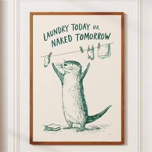Peut inclure: Une affiche encadrée présentant un dessin au trait vert d'une loutre debout, les bras levés. Au-dessus de la loutre, une corde à linge porte une chaussette et un vêtement avec des pinces. Le texte indique : LAUNDRY TODAY OR NAKED TOMORROW.