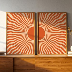 Könnte beinhalten: Zwei rechteckige gerahmte Wandbilder mit einem symmetrischen Sonnenstrahlen-Motiv. Jeder Druck zeigt einen massiven orangefarbenen kreisförmigen Mittelpunkt mit wellenförmigen orangefarbenen und cremefarbenen Linien.