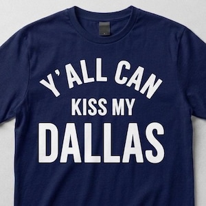 Op de afbeelding: Een marineblauw t-shirt met korte mouwen en de witte tekst Y'ALL CAN KISS MY DALLAS in een vet, schreefloos lettertype op de borst. Het shirt heeft een ronde hals en een standaard pasvorm, met een leeg label zichtbaar in de kraag.