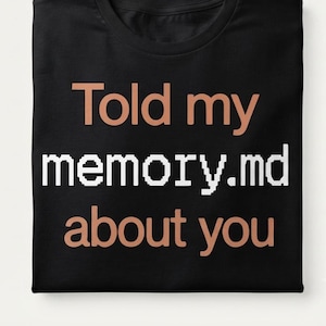May include: A black t-shirt featuring the text "Told my" in a light peach colour, followed by "memory.md" in a white pixelated font, and "about you" in a light peach colour. The shirt is folded neatly, displaying the graphic centred on the front.