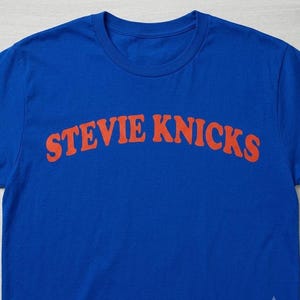 May include: A bright blue short-sleeved t-shirt featuring the text STEVIE KNICKS printed in a bold, arched orange font across the chest. The garment has a classic crew neckline and is made of a solid-colored fabric with a smooth texture.